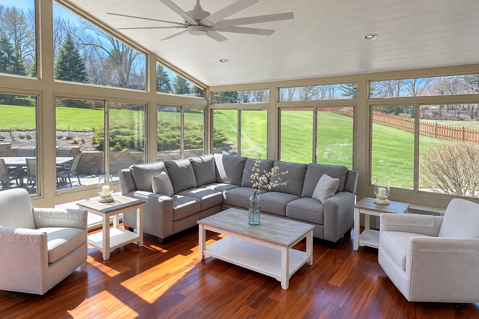 Sunroom with large windows, a gray sectional, white armchairs, and polished wood flooring beside a green lawn.