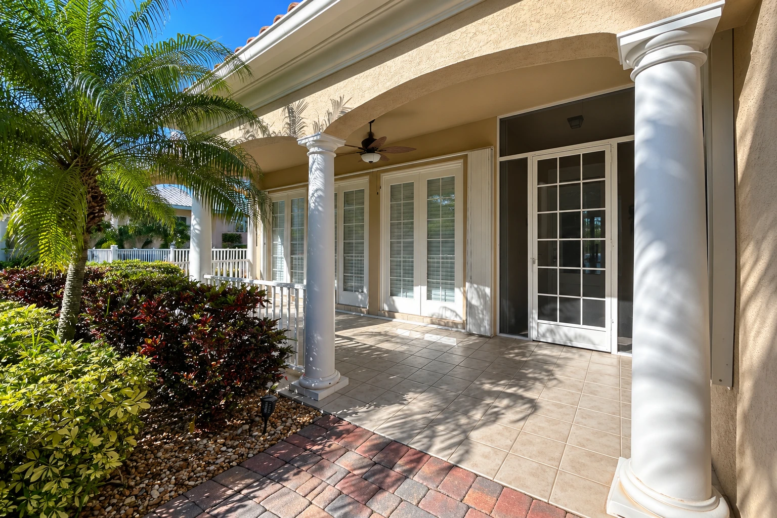 Covered patio with tiled flooring, white columns, and a screened door beside tropical landscaping.