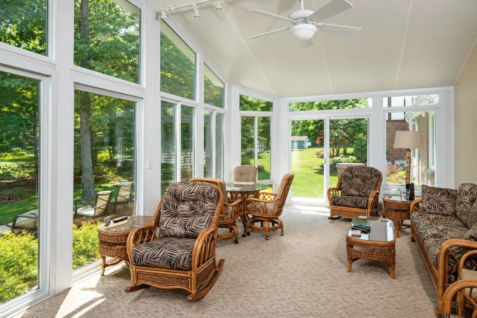 Bright sunroom with white framing, expansive windows, wicker seating, and a view of a leafy lawn.