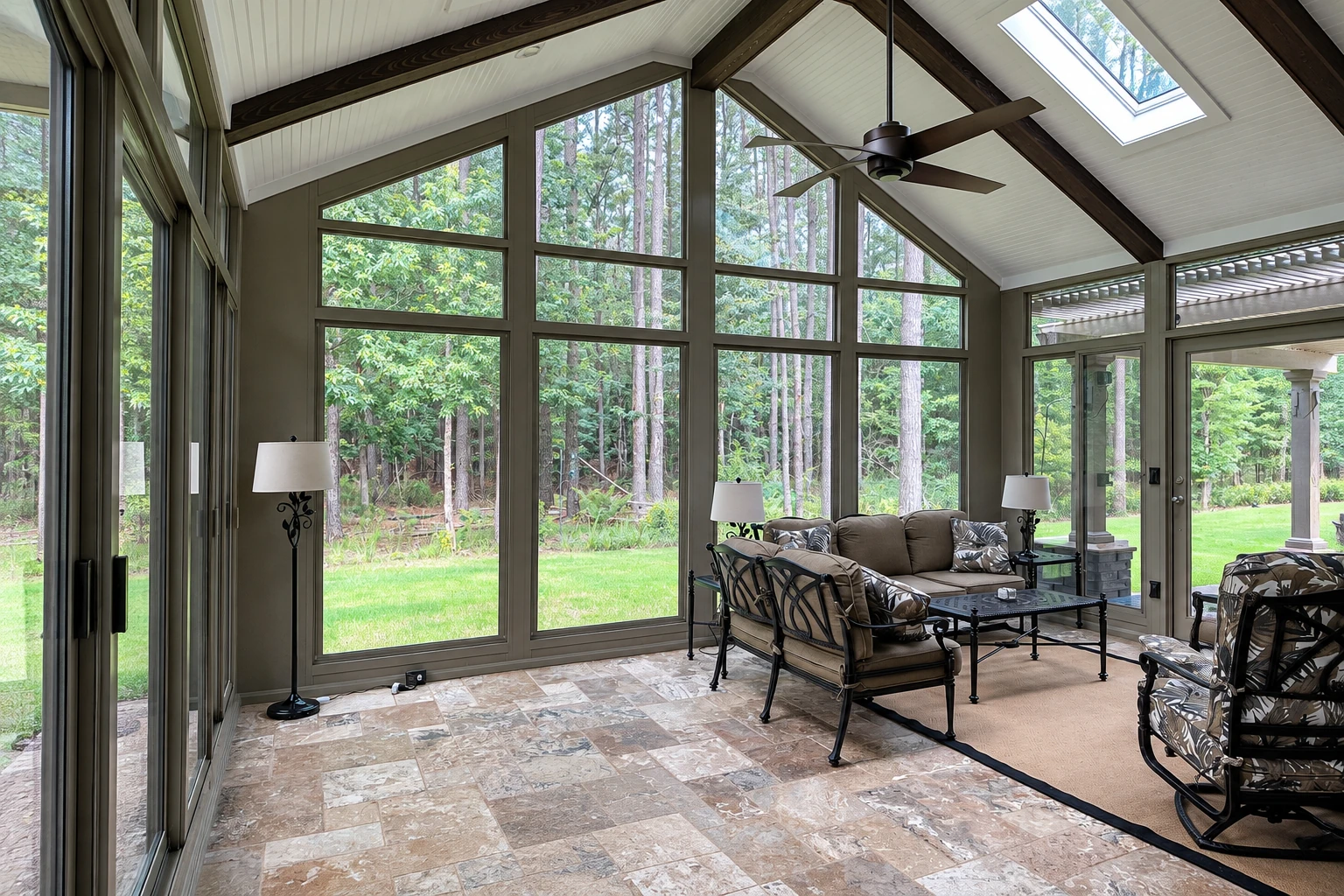 Sunroom with tall windows, a vaulted ceiling, skylight and dark-framed seating overlooks a wooded yard.