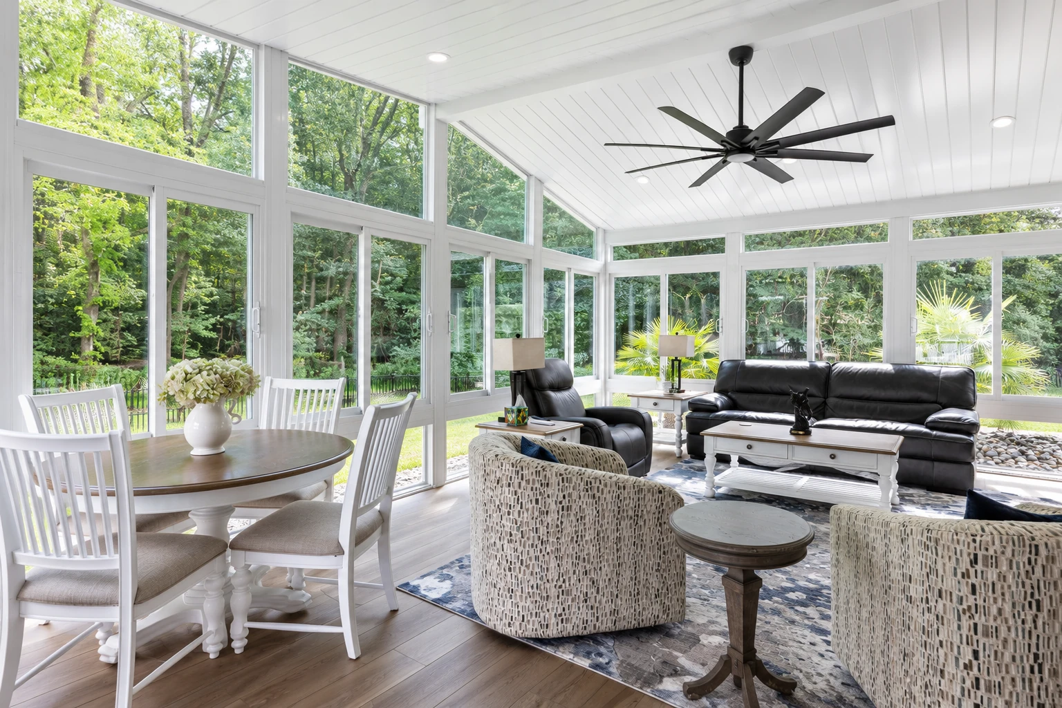 A bright sunroom with white framing and tall windows contains a dining table, armchairs, and a black sofa beneath a ceiling fan.