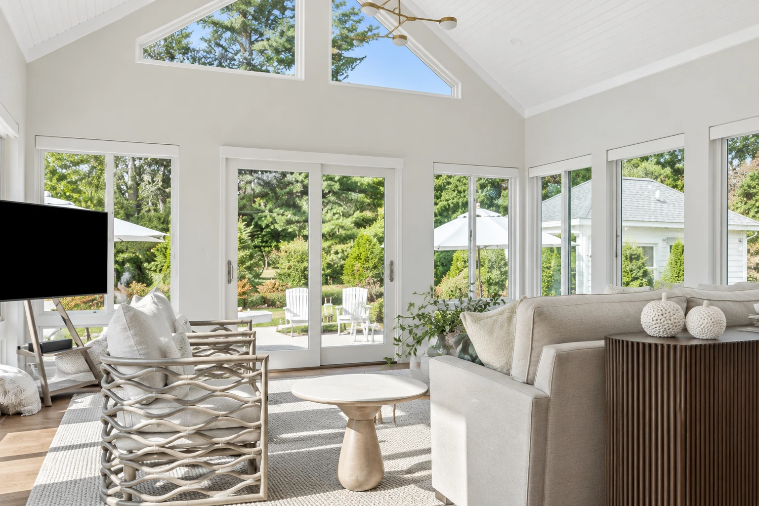 A white sunroom with a vaulted ceiling and high windows is furnished with a sofa and wicker chairs overlooking a garden.